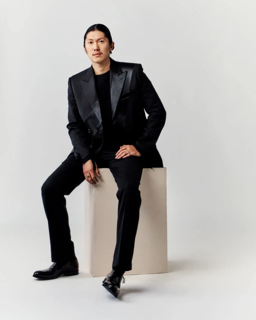 Stylish Asian man in a black suit sitting on a white block in a studio. He is wearing a ring and black leather shoes.
