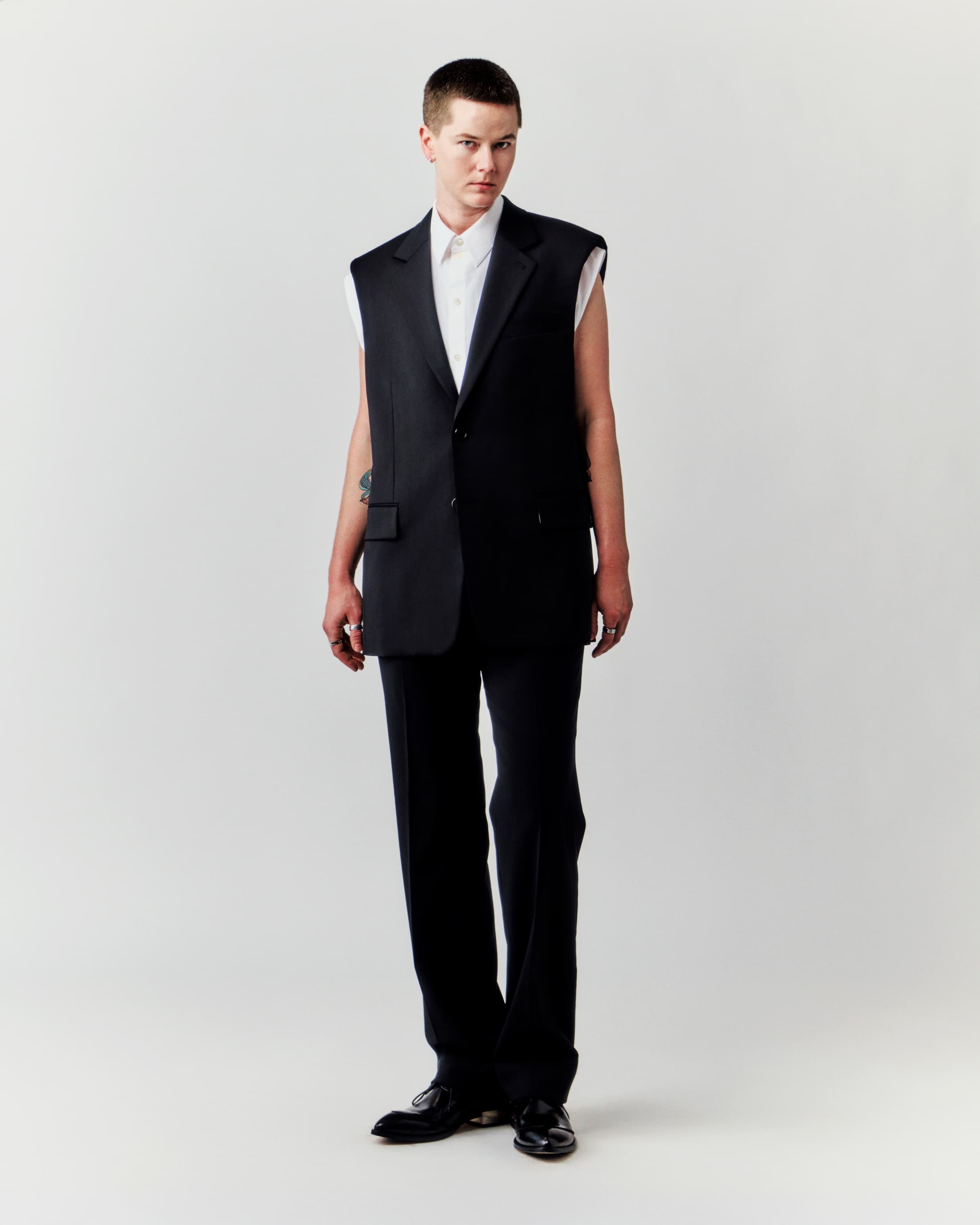 Studio shot of a young adult male in a sleeveless black blazer and trousers against a clean white backdrop, stylish and modern.