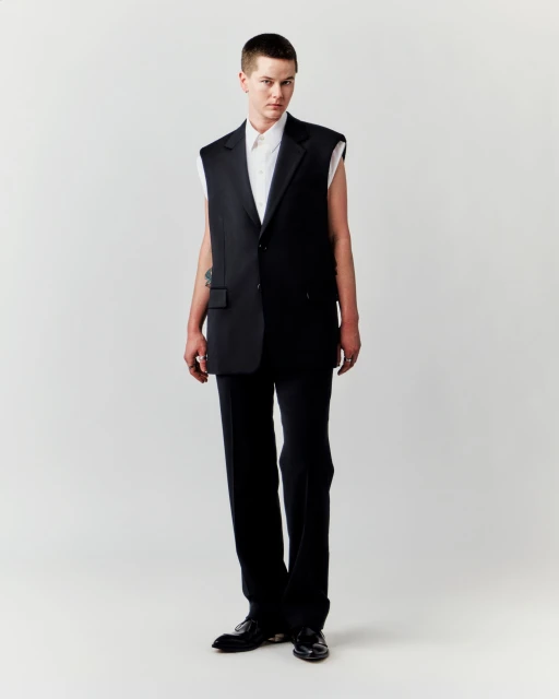 Studio shot of a young adult male in a sleeveless black blazer and trousers against a clean white backdrop, stylish and modern.