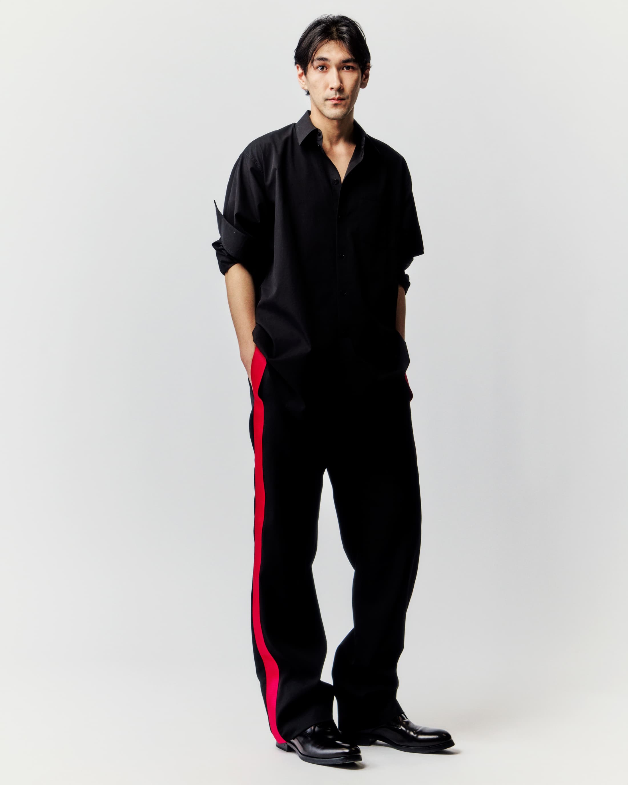 Studio shot of a young adult man in a loose black shirt and trousers with a red stripe, complemented by black shoes on a light backdrop.