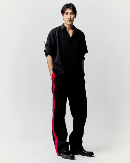 Studio shot of a young adult man in a loose black shirt and trousers with a red stripe, complemented by black shoes on a light backdrop.