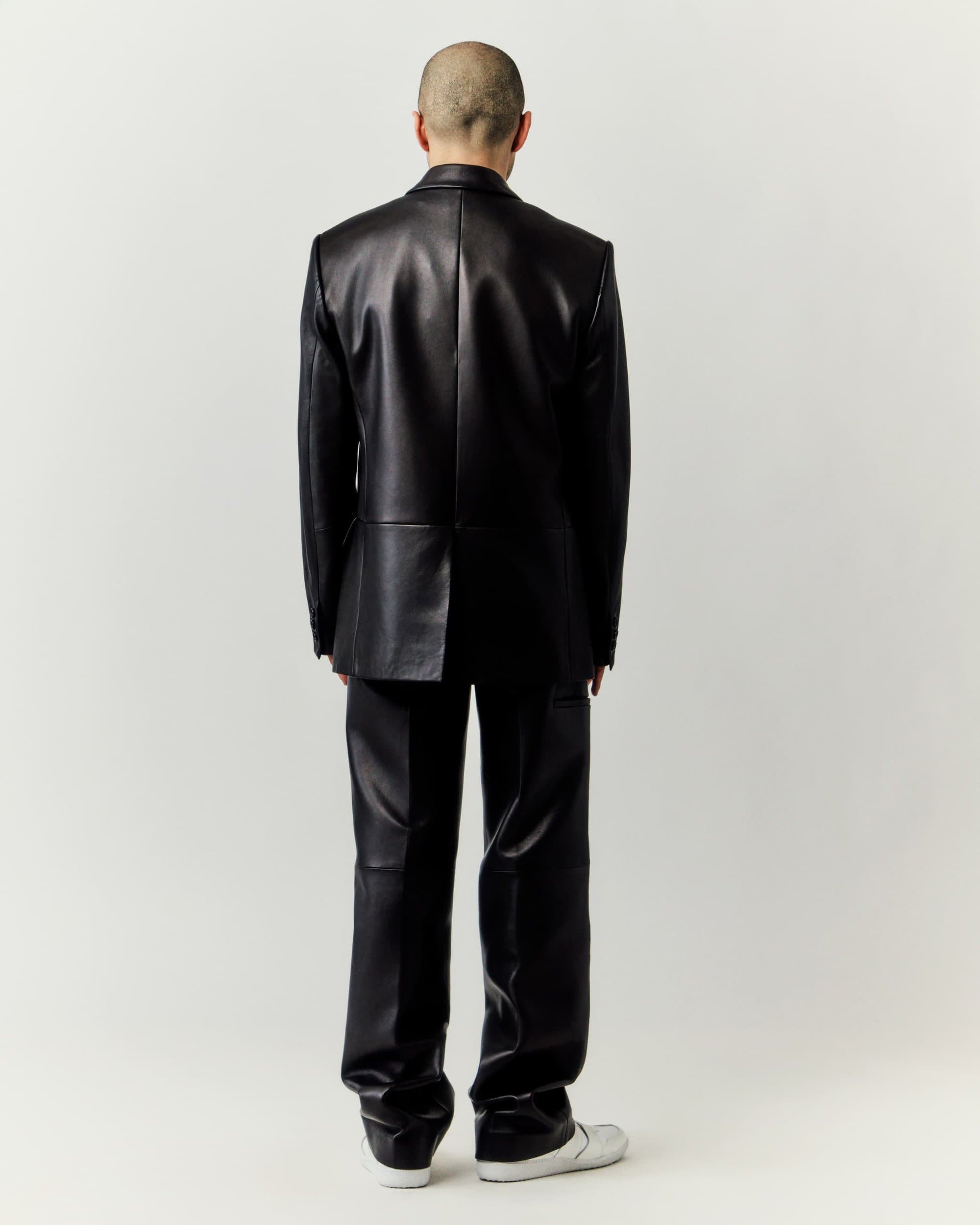 An adult male stands with his back to the camera wearing a shiny black leather suit with sneakers against a neutral background.