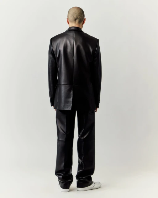 An adult male stands with his back to the camera wearing a shiny black leather suit with sneakers against a neutral background.