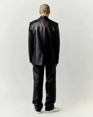 An adult male stands with his back to the camera wearing a shiny black leather suit with sneakers against a neutral background.