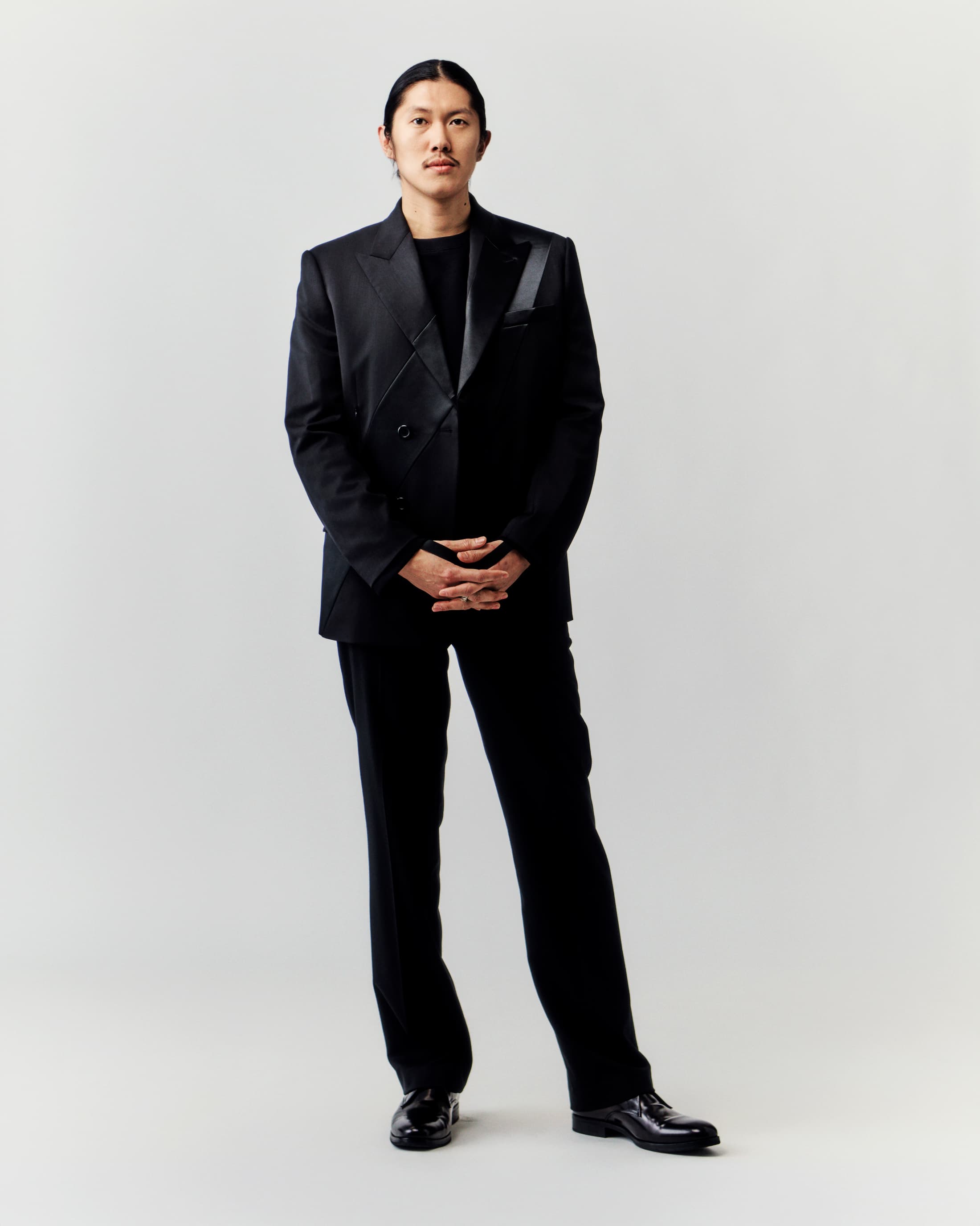 Full-length portrait of a stylish adult male in a black suit posing indoors against a neutral background, showcasing fashion and professional style.