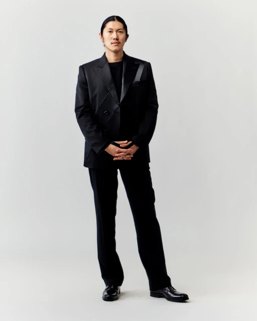 Full-length portrait of a stylish adult male in a black suit posing indoors against a neutral background, showcasing fashion and professional style.