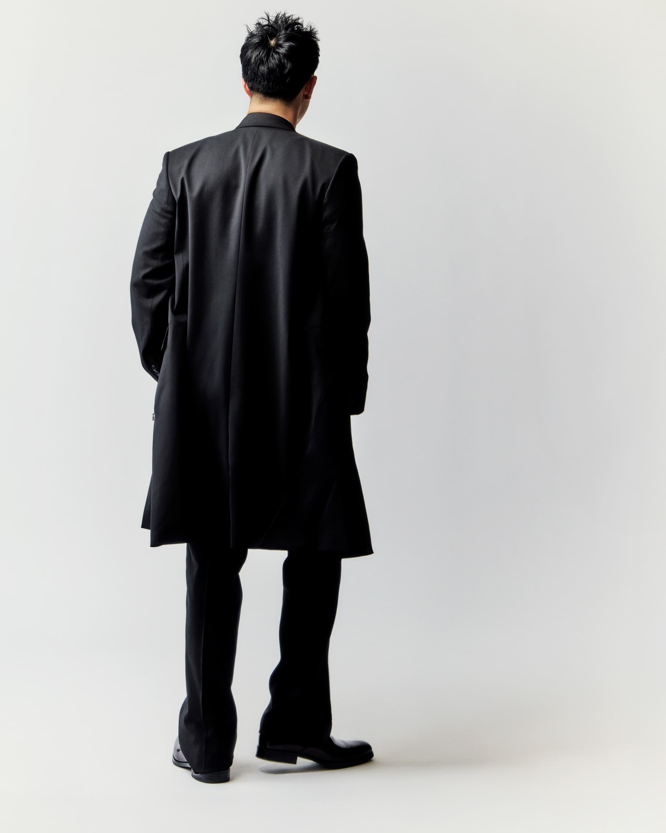 An adult male stands with his back to the camera, showcasing a long black coat, dark pants, and dress shoes against a clean white backdrop.