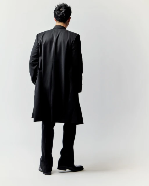 An adult male stands with his back to the camera, showcasing a long black coat, dark pants, and dress shoes against a clean white backdrop.