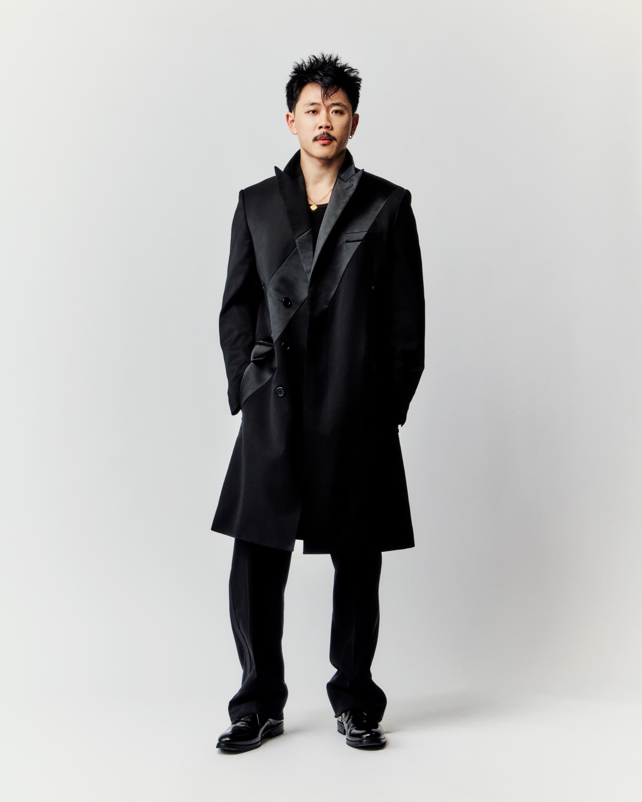 A man stands wearing a stylish black coat and pants, set against a clean, light background in a full-length portrait shot.