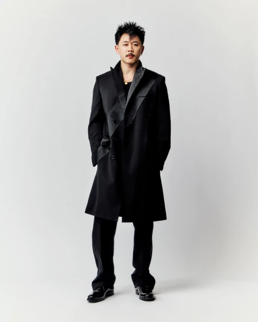 A man stands wearing a stylish black coat and pants, set against a clean, light background in a full-length portrait shot.