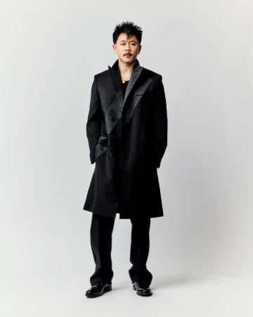 A man stands wearing a stylish black coat and pants, set against a clean, light background in a full-length portrait shot.