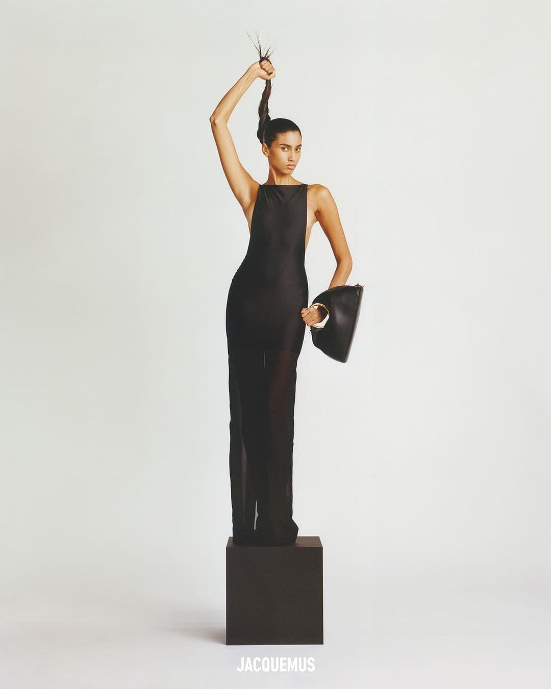 Fashion shot of a woman in a black dress holding a purse, standing on a black cube against a white backdrop in a studio.