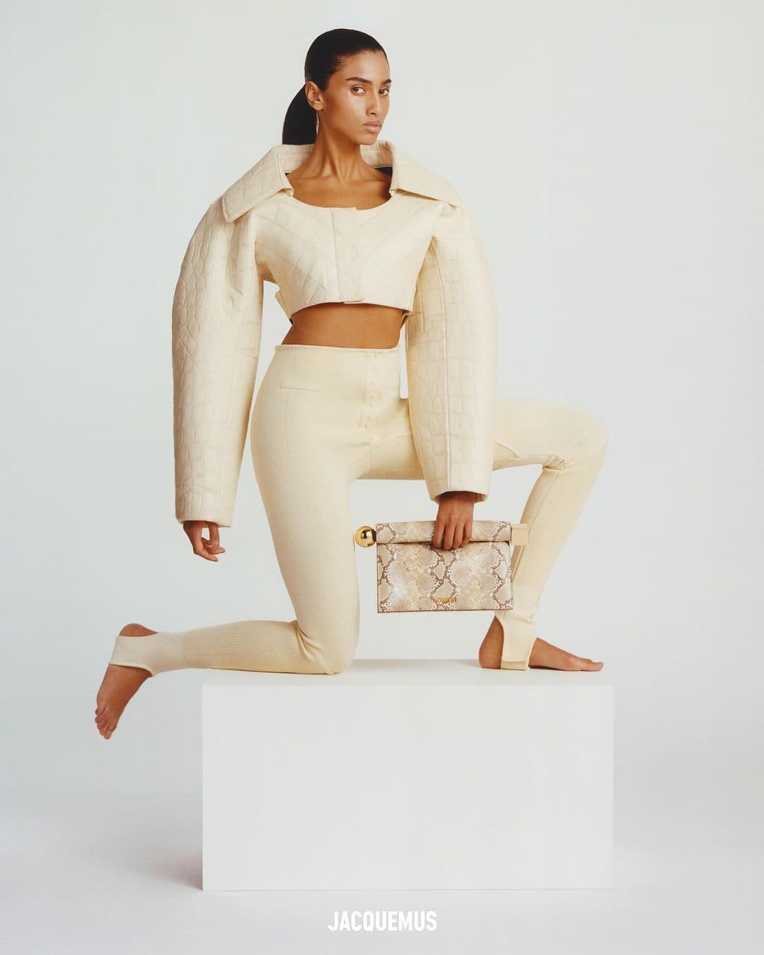 A stylish adult female with tanned skin poses in a cream-colored, quilted two-piece outfit against a white backdrop, holding a snakeskin clutch.