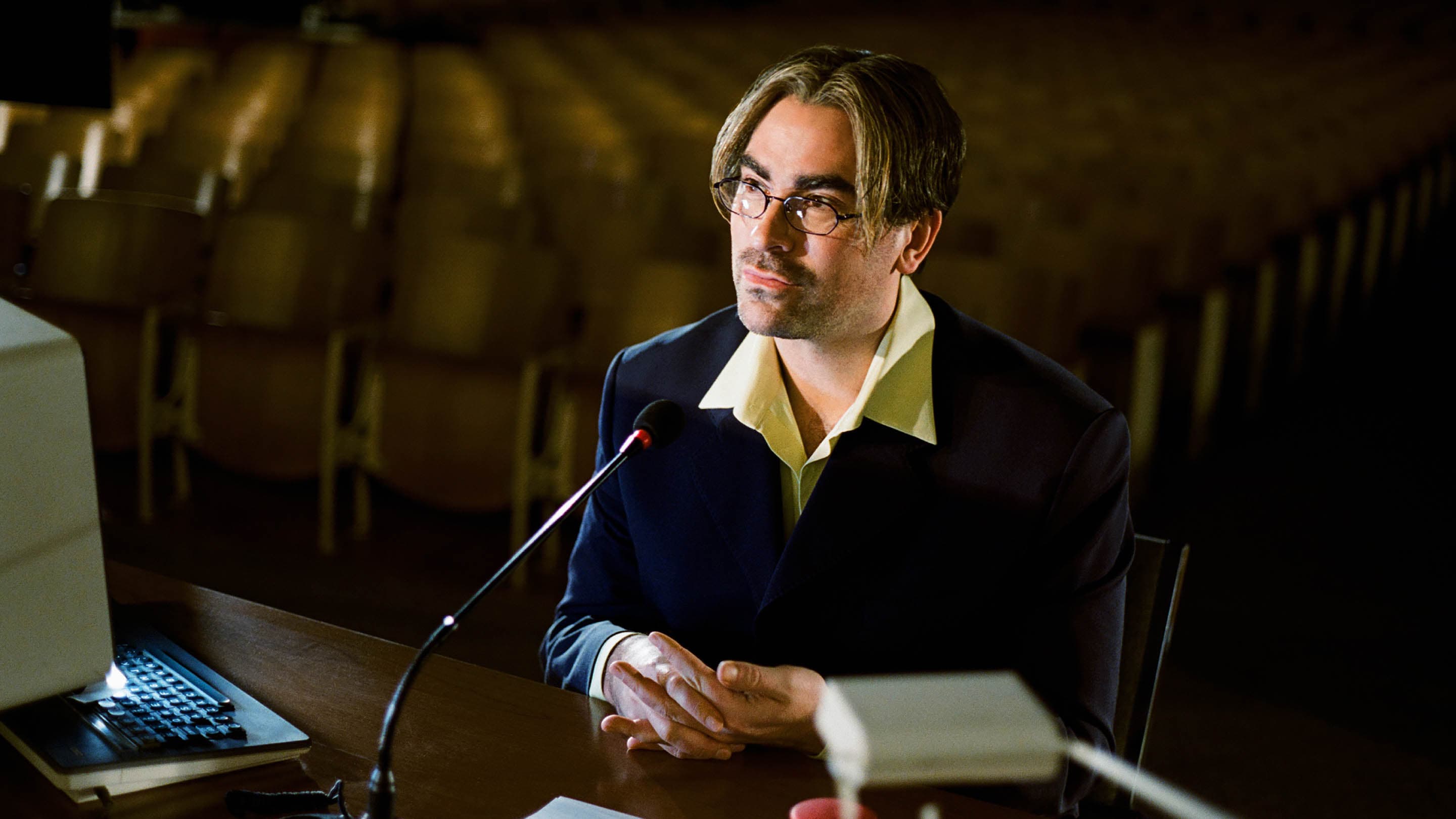 Adult male presenter wearing glasses and a dark suit at a wooden desk with a microphone and laptop in an auditorium setting.