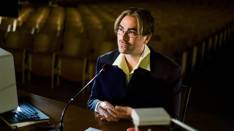Adult male presenter wearing glasses and a dark suit at a wooden desk with a microphone and laptop in an auditorium setting.