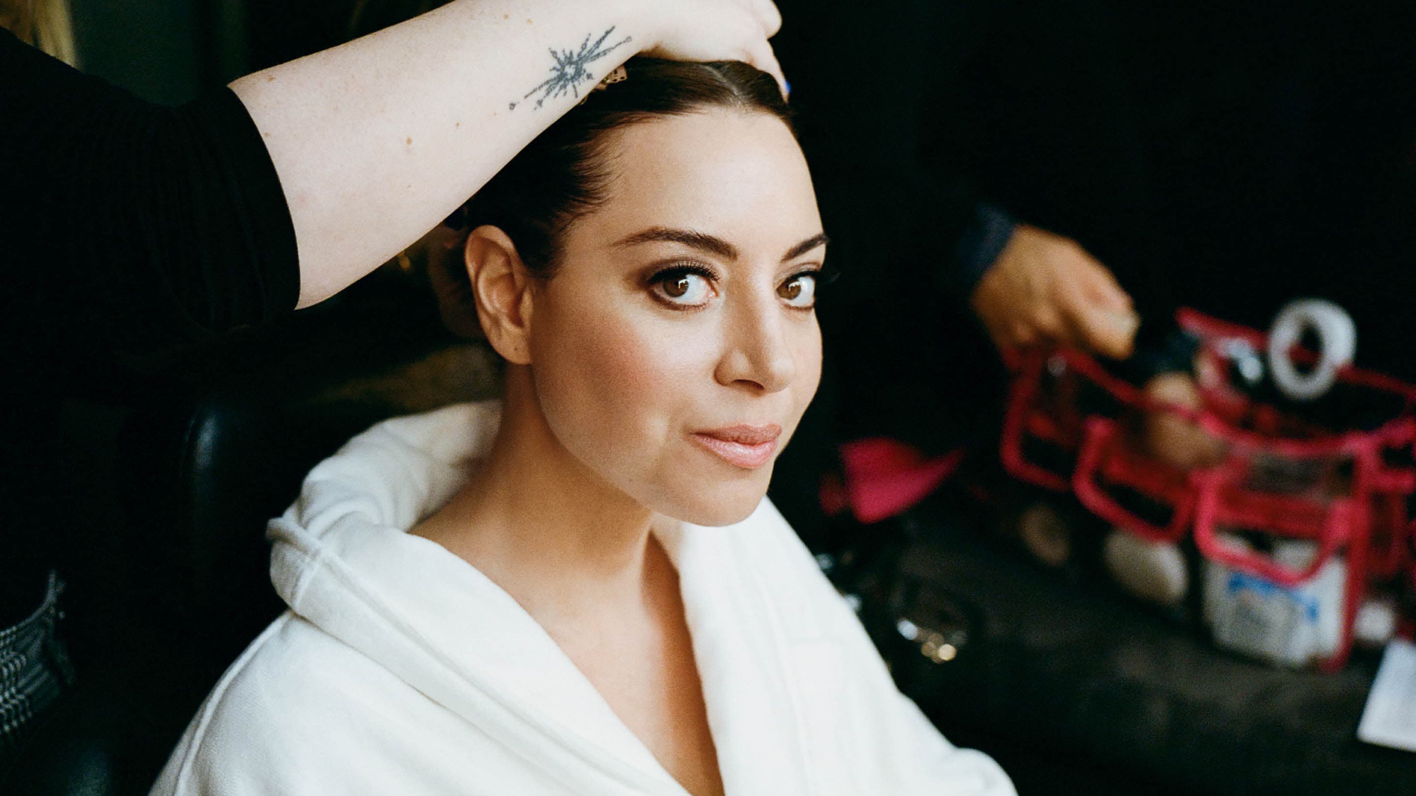 Close-up of a woman with fair skin and dark hair having her hair styled, wearing a white robe in a softly lit indoor setting.