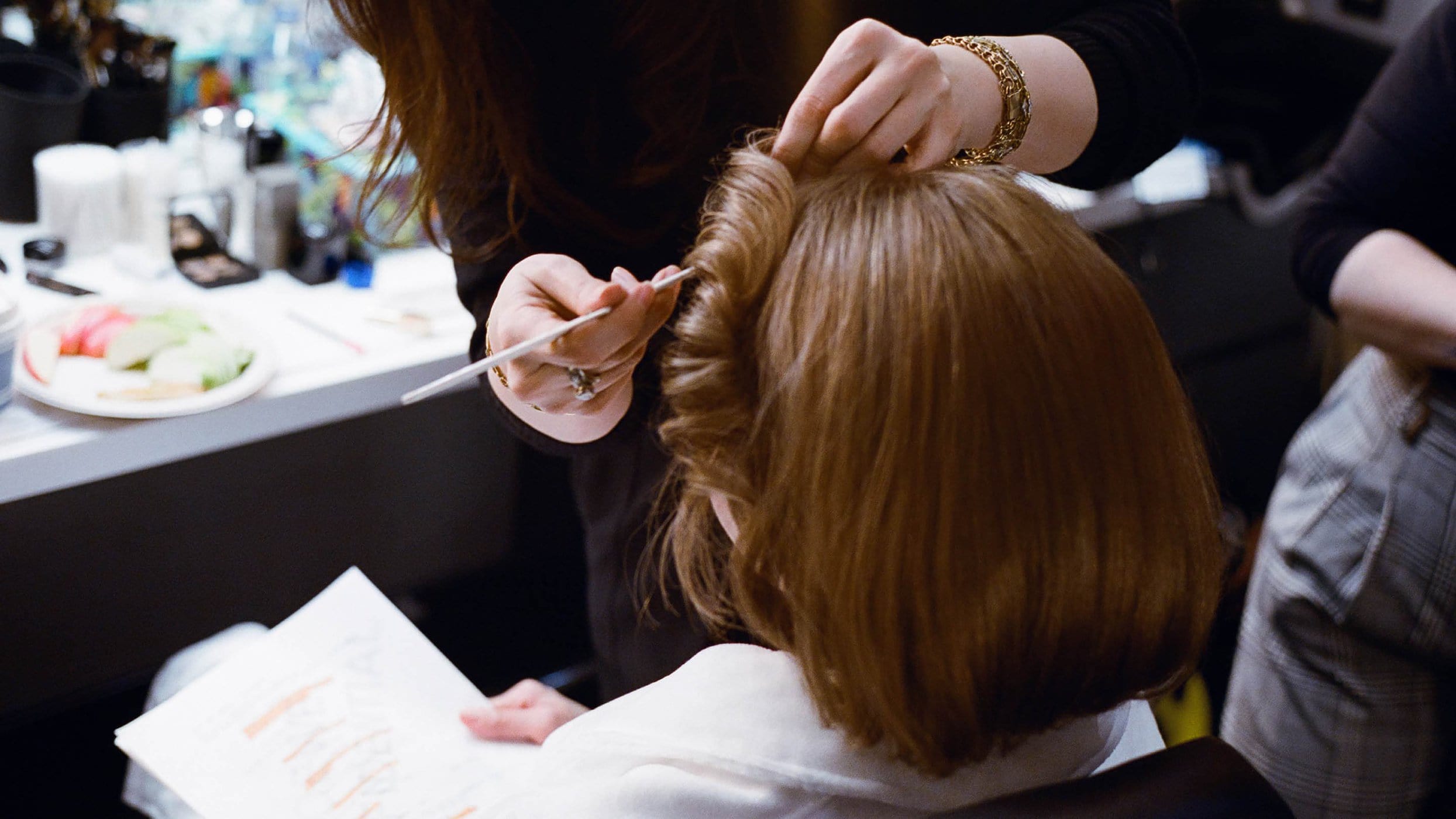 Woman getting her hair styled by a hairstylist using a thin tool, at a salon or backstage, with a light and bright atmosphere.
