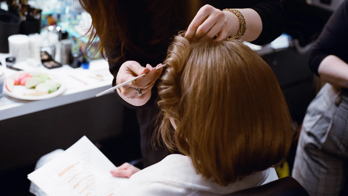 Woman getting her hair styled by a hairstylist using a thin tool, at a salon or backstage, with a light and bright atmosphere.