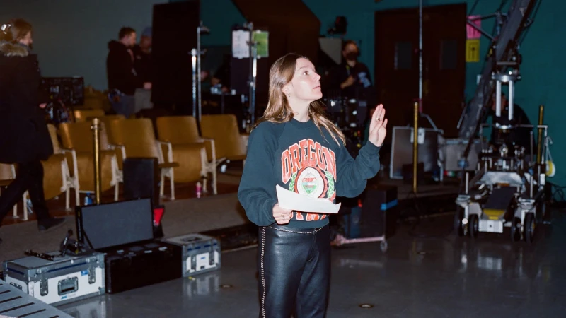 A woman in a sweatshirt and leather pants directs a film crew on a set with theater seating and equipment, in a medium indoor shot.