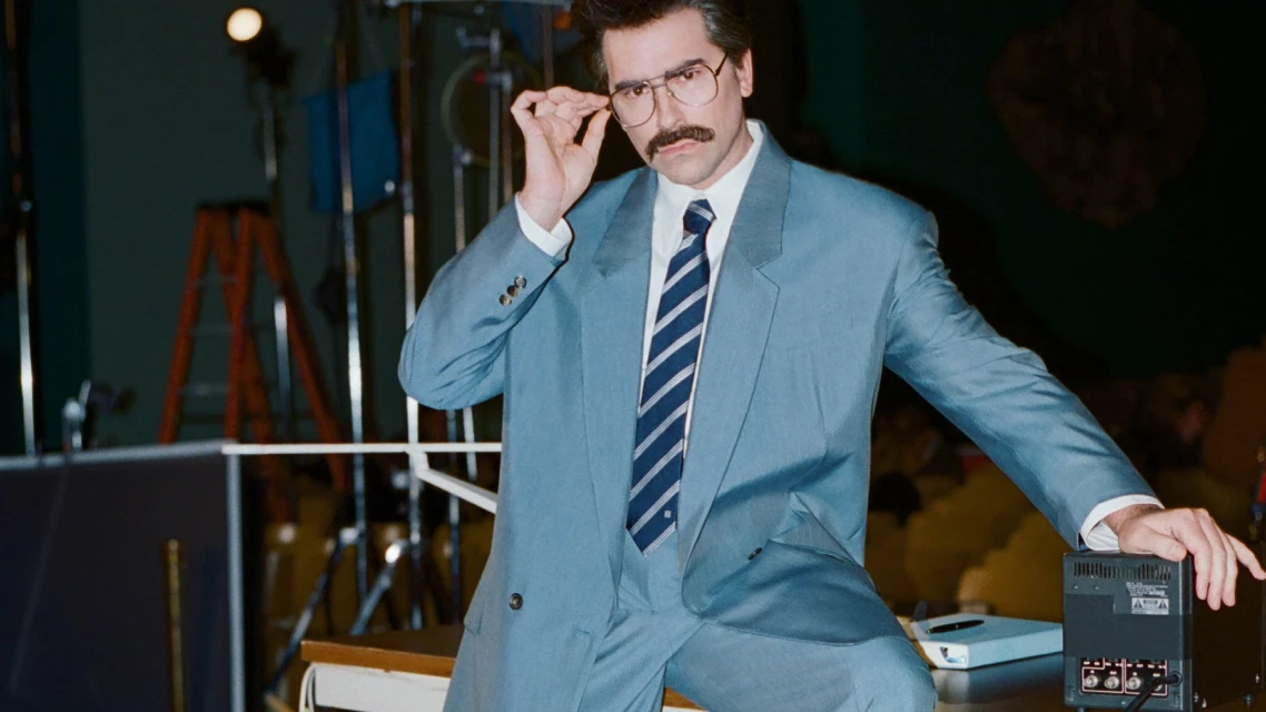 Adult male in a light blue suit adjusts his glasses while sitting on a desk in a photo studio with lighting equipment visible.