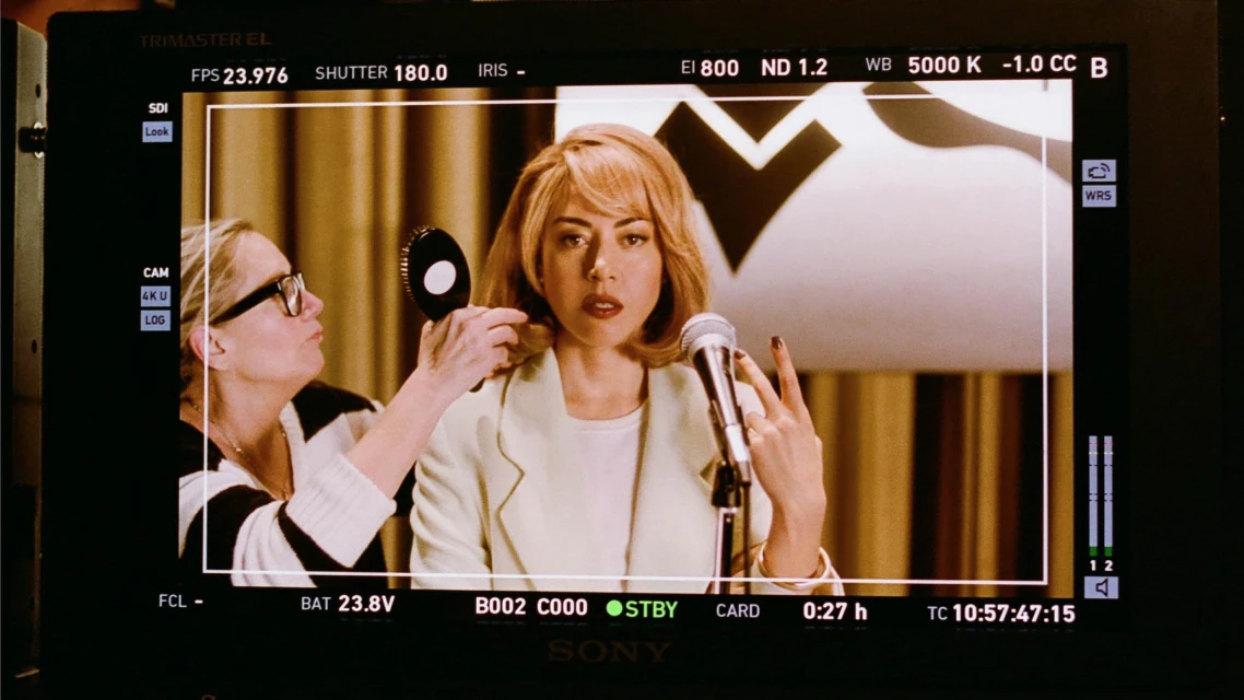 Aubrey Plaza as April Ludgate on Parks and Recreation being prepped with hair and makeup before speaking into a microphone.