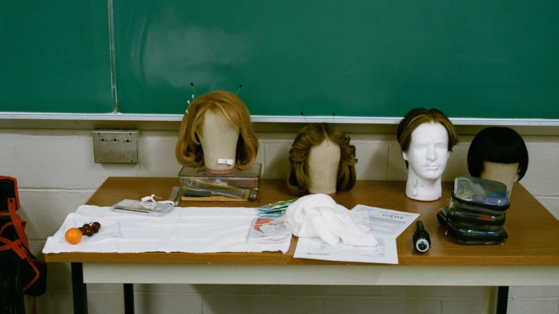 Assortment of mannequin heads with wigs, along with various objects and props, displayed on a table in front of a green chalkboard.