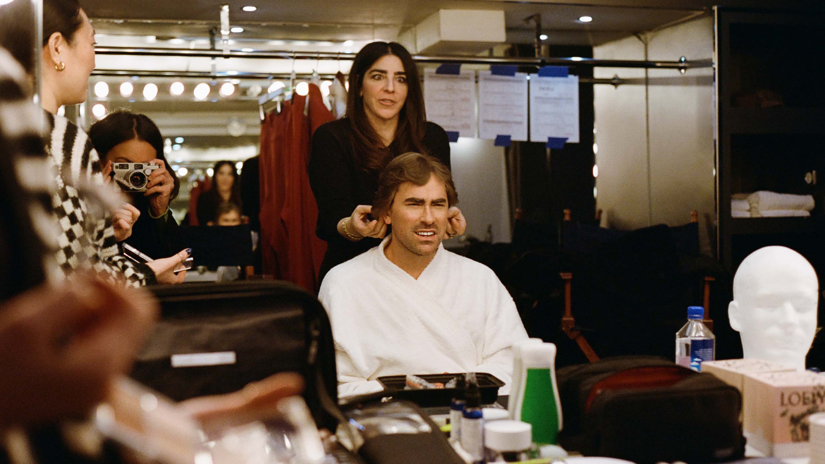 Man getting hair styled in dressing room mirror with makeup, hair products, and other stylists visible in the background.