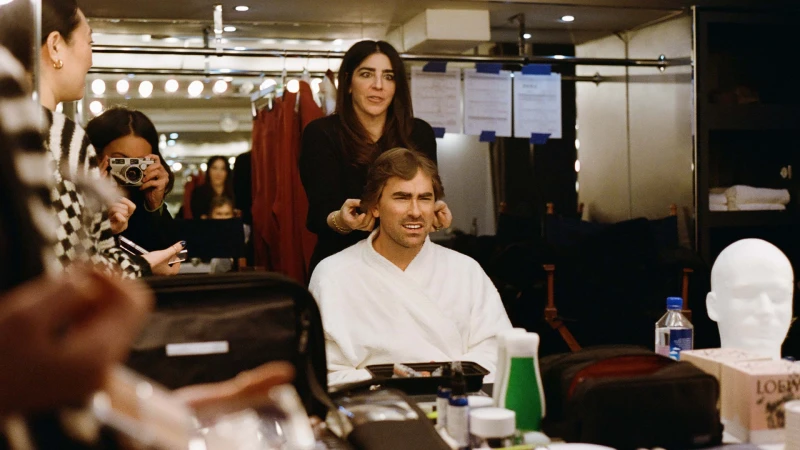 Man getting hair styled in dressing room mirror with makeup, hair products, and other stylists visible in the background.