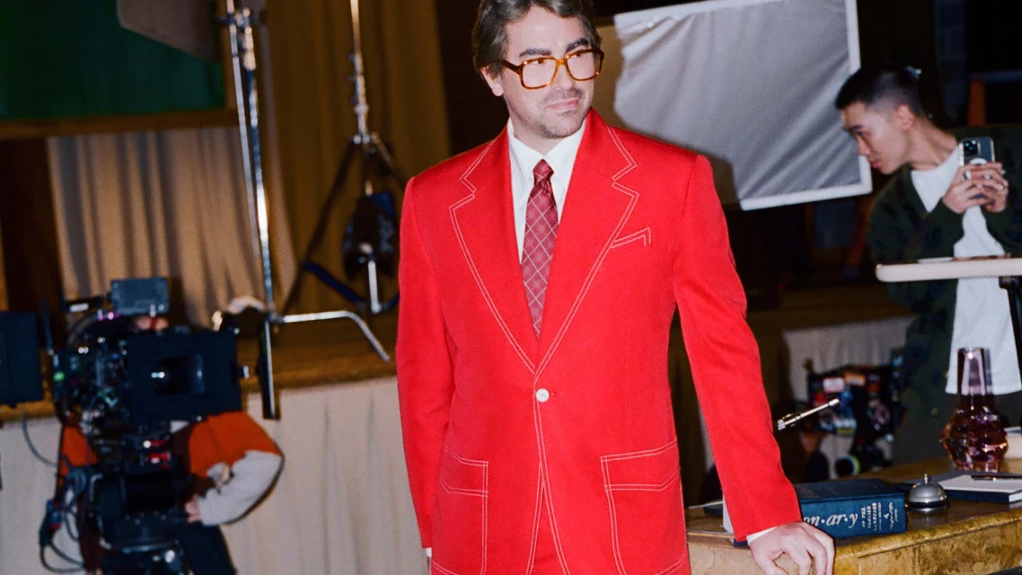 Man in a bright red suit stands in front of a camera and film crew. The setting appears to be in a photo studio for filming.