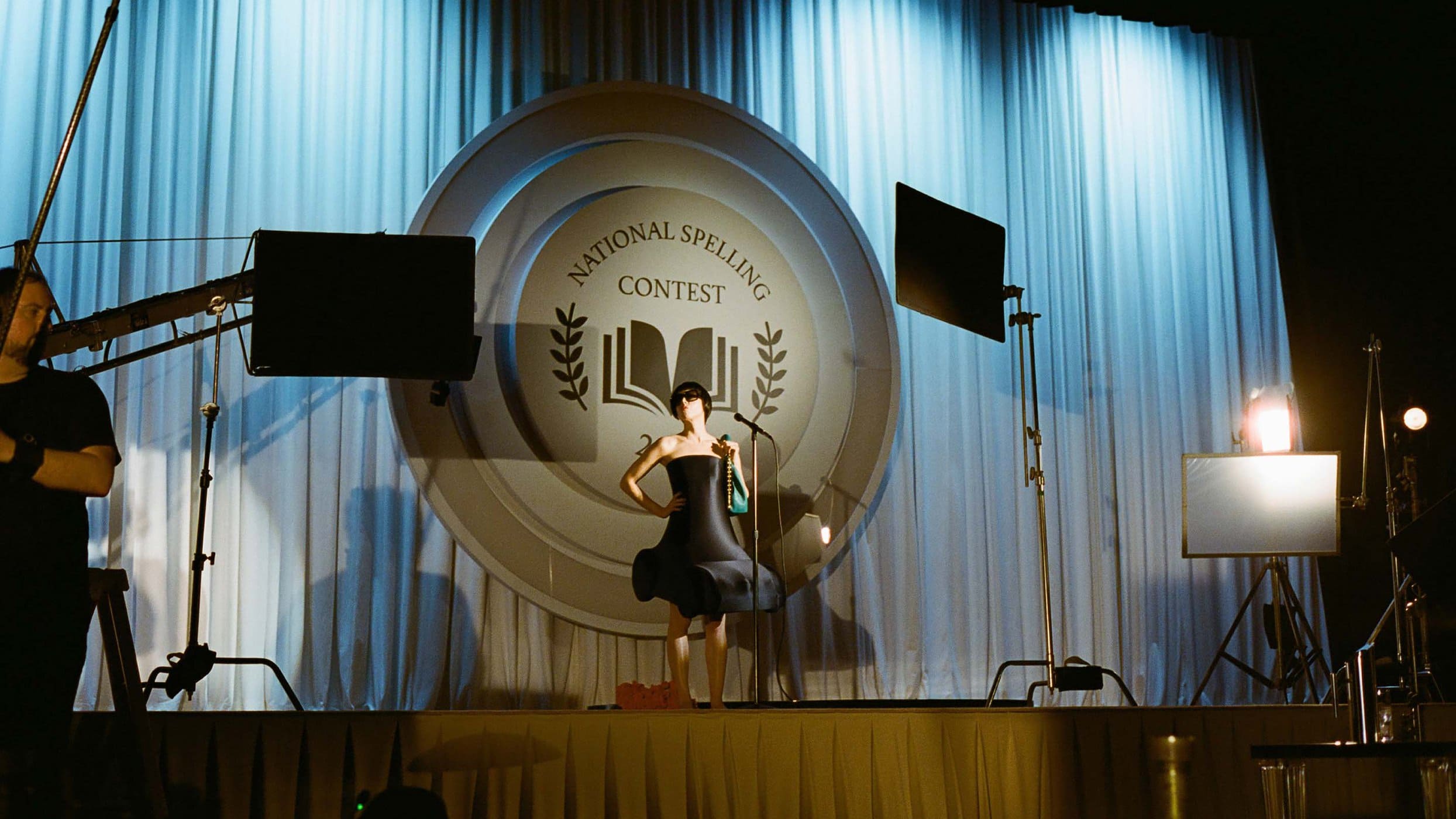 Woman in black dress posing on stage with National Spelling Contest backdrop, stage lighting, and a camera operator visible.