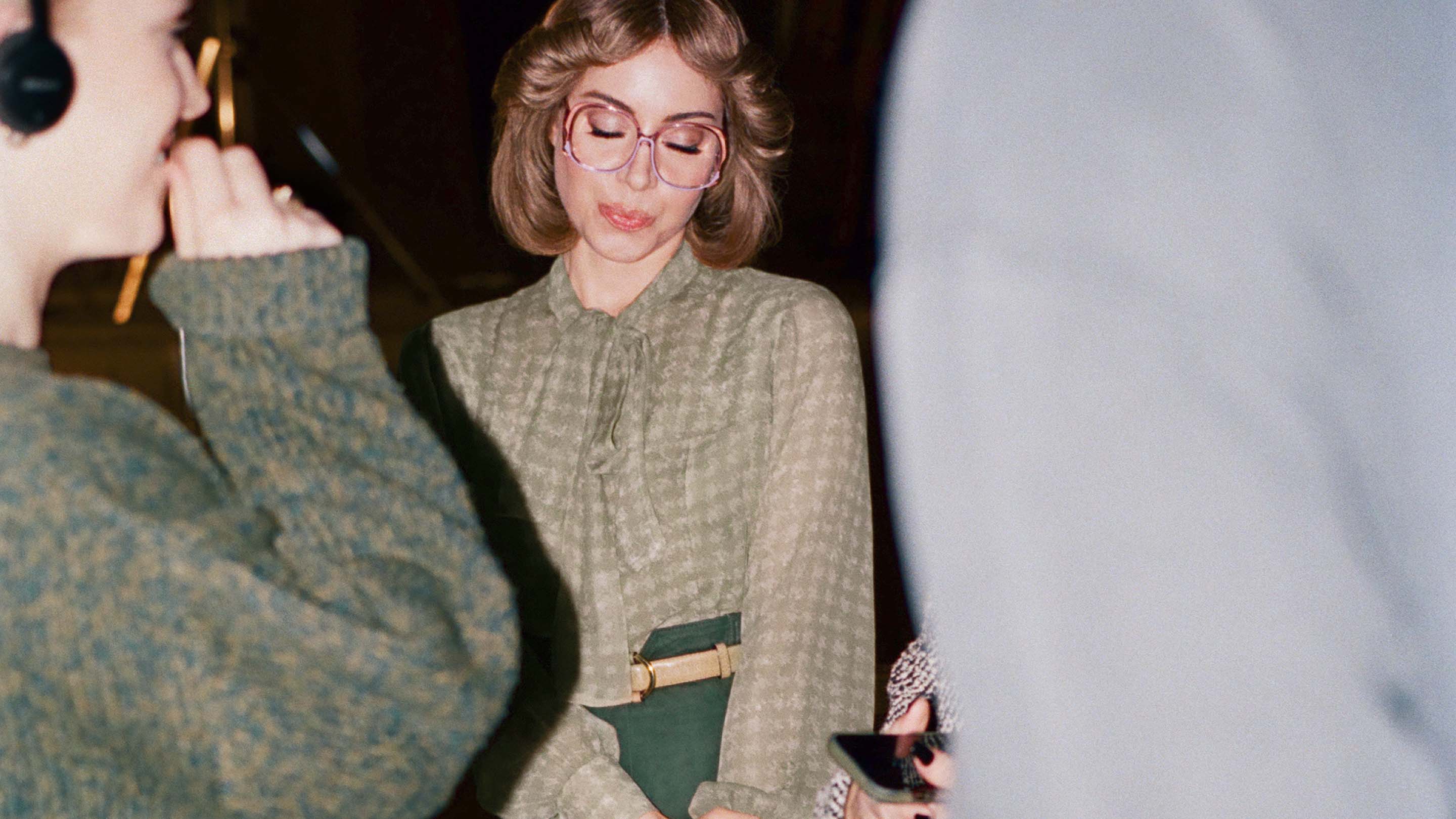 Woman in patterned blouse and glasses standing indoors near another woman with headphones in a fashion setting with warm lighting.