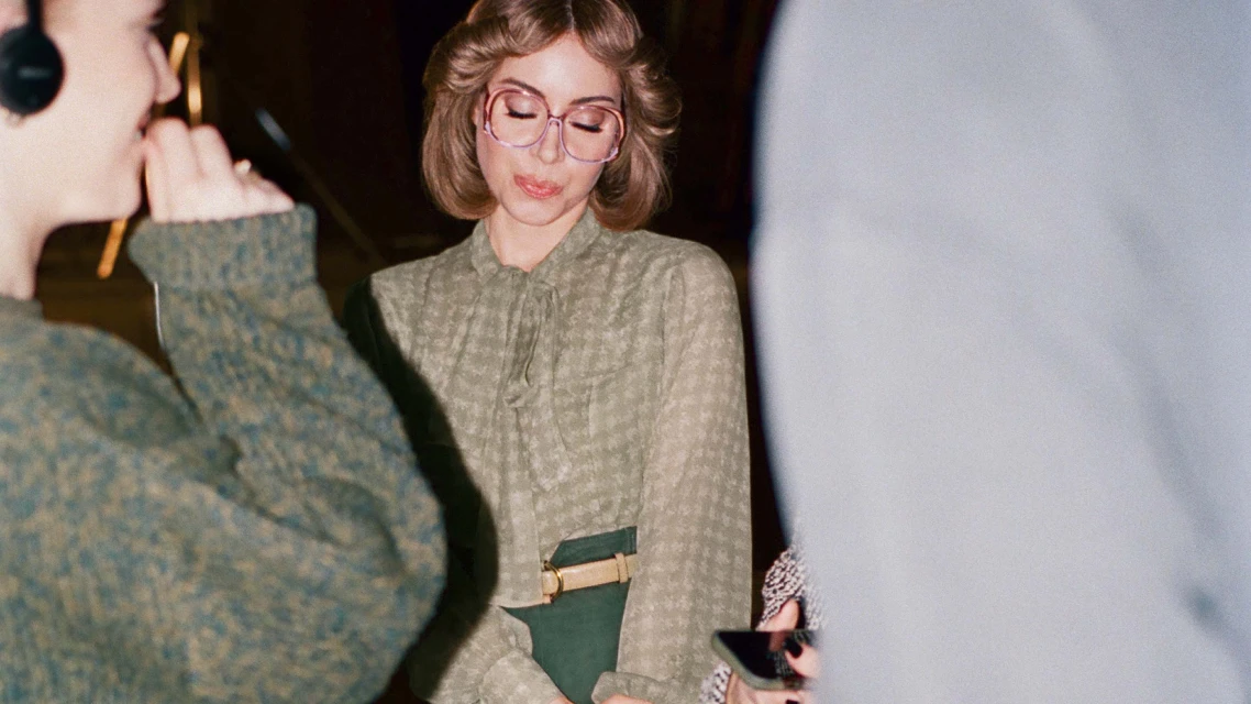 Woman in patterned blouse and glasses standing indoors near another woman with headphones in a fashion setting with warm lighting.