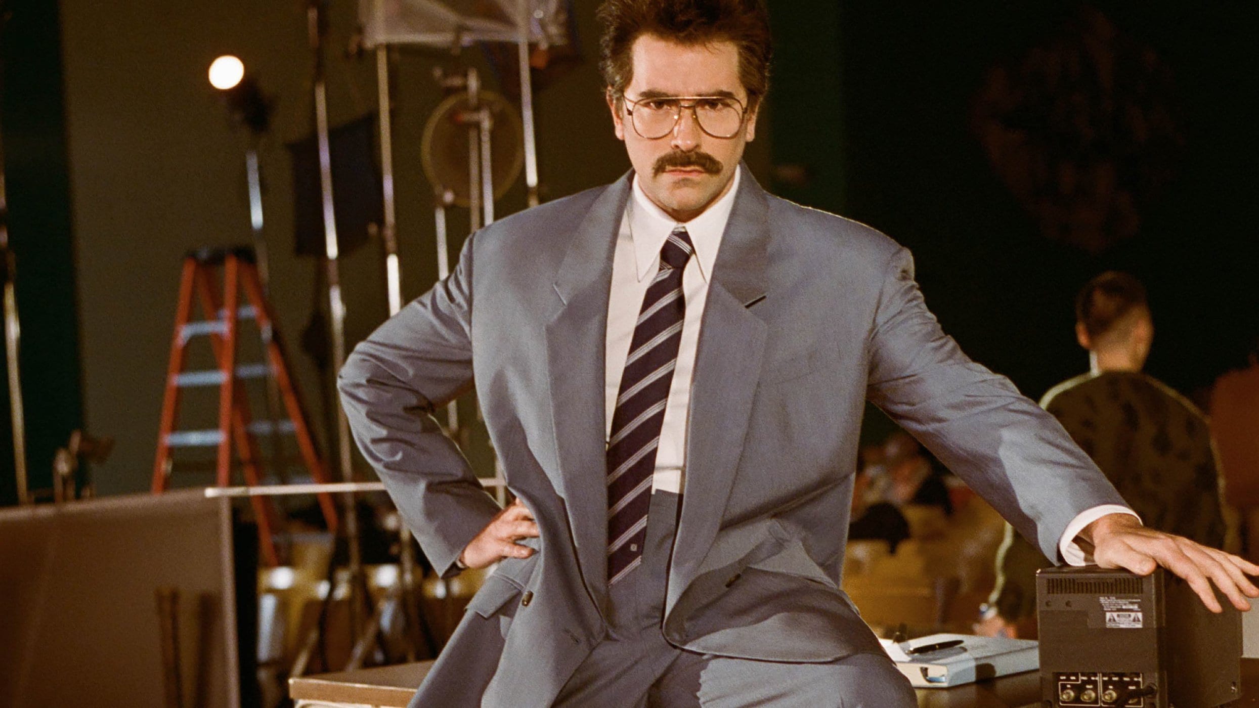 Adult male in a grey suit with a mustache, posing in what looks like a studio, sitting on desk with old electronics and equipment.