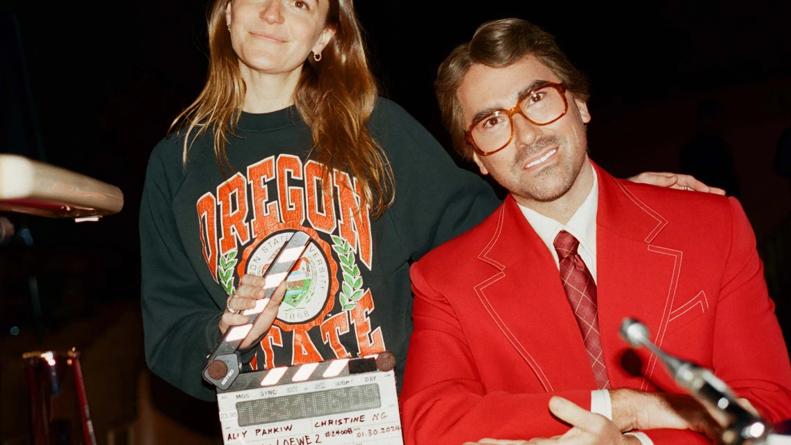 Two people pose indoors: a person in a red suit and glasses, and another in a sweatshirt holding a film slate.
