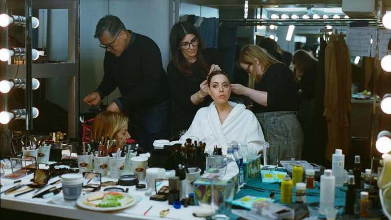 Woman in a white bathrobe getting her hair and makeup done by a team of stylists in a dressing room, with visible makeup products.