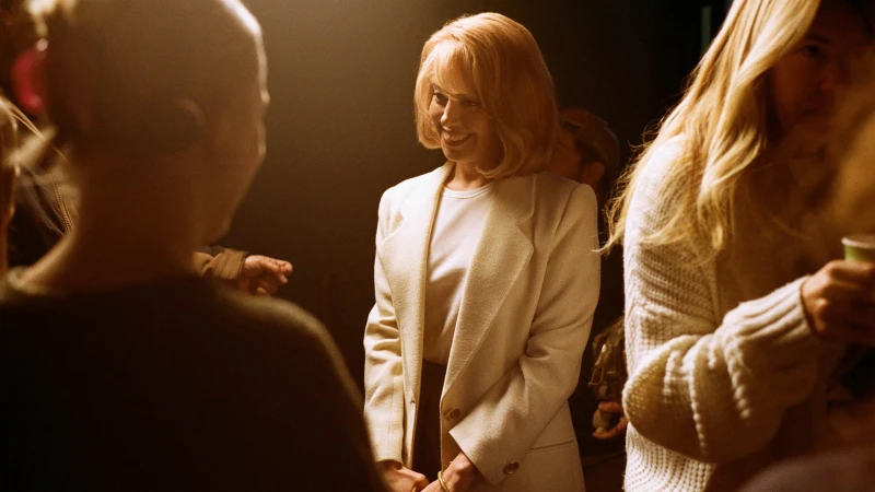 A blonde woman in a white blazer and t-shirt smiles while standing in a dimly lit, crowded room with warm spotlighting.