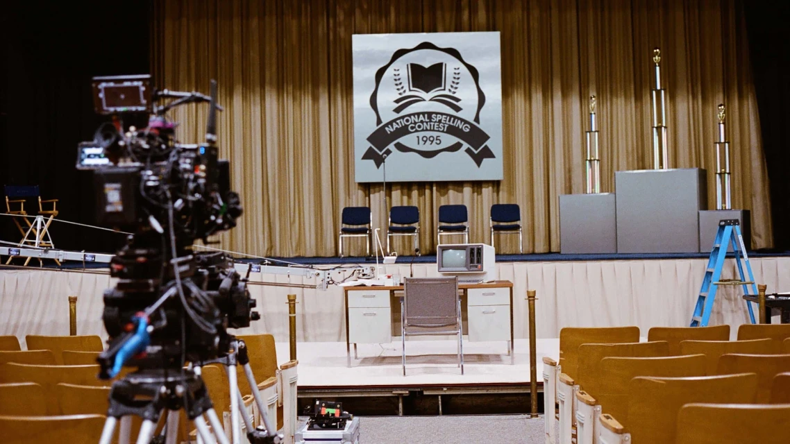 Empty theater with a large television camera in foreground, stage with a desk, podiums, chairs, and the National Spelling Bee logo.