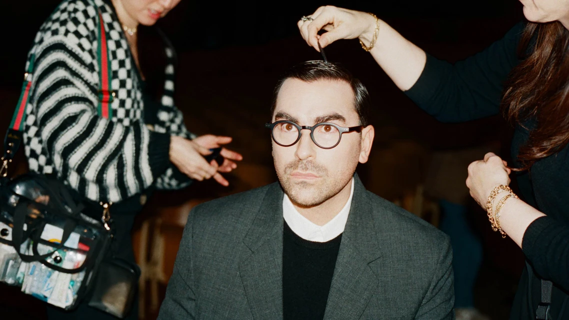 Man getting hair styled by two people in an indoor setting, wearing a gray suit and glasses. Focused portrait with dark background.