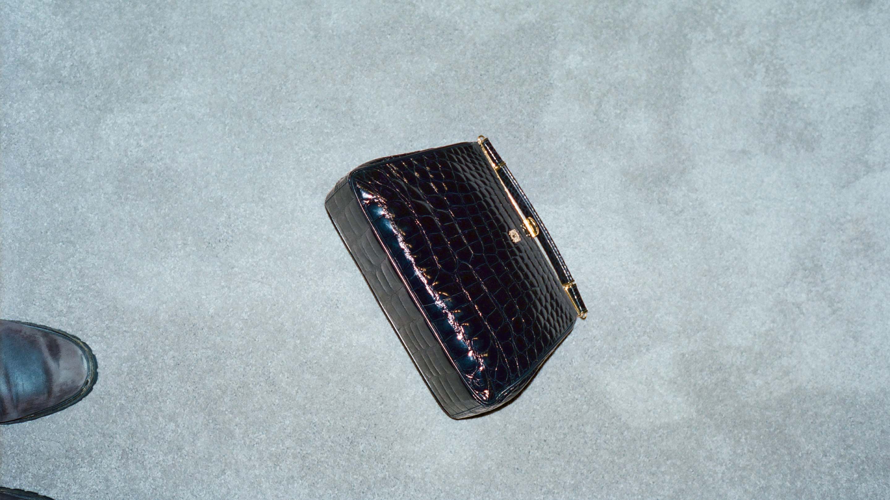 Elevated shot of a black alligator-skin handbag with gold hardware, lying on a textured light gray surface with a shoe in the corner.