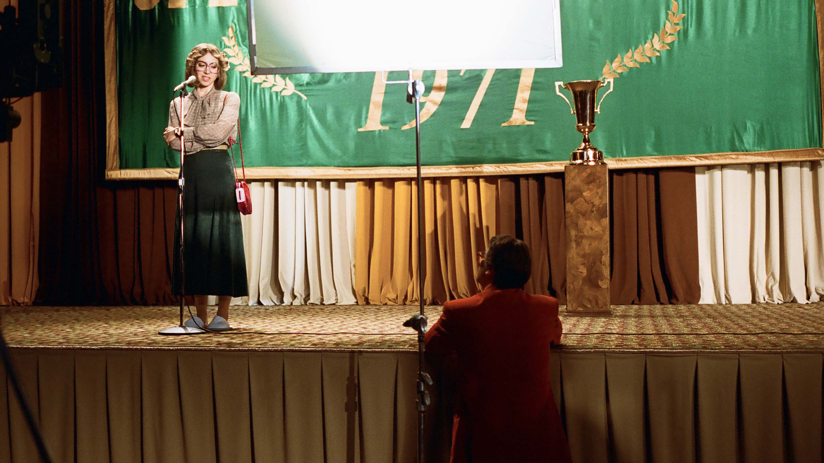 Woman giving a speech on stage in front of a banner that says 1971, with a golden trophy to her right and a seated man in the foreground.