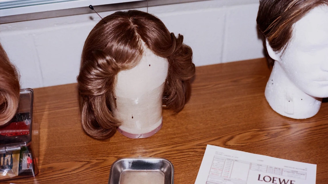Wig on a mannequin head on a table with styling tools and a Loewe document. Light brown wig styled in waves.