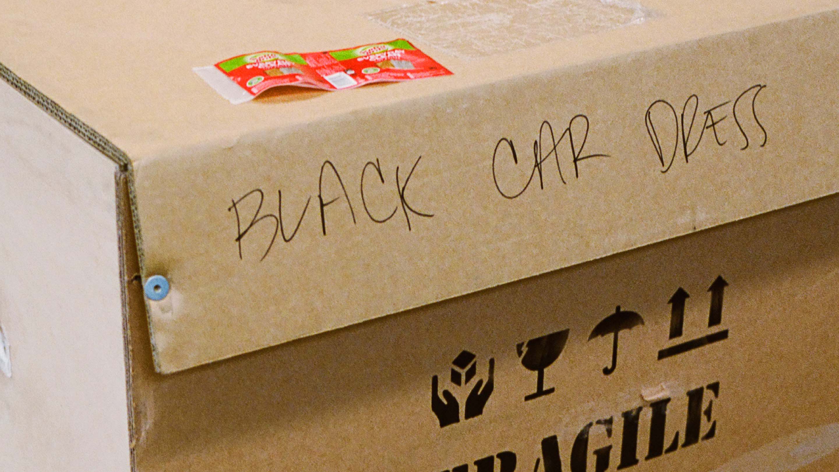 Close-up of a brown cardboard shipping box with handwritten text that says 'BLACK CAR DRESS' and package handling symbols.