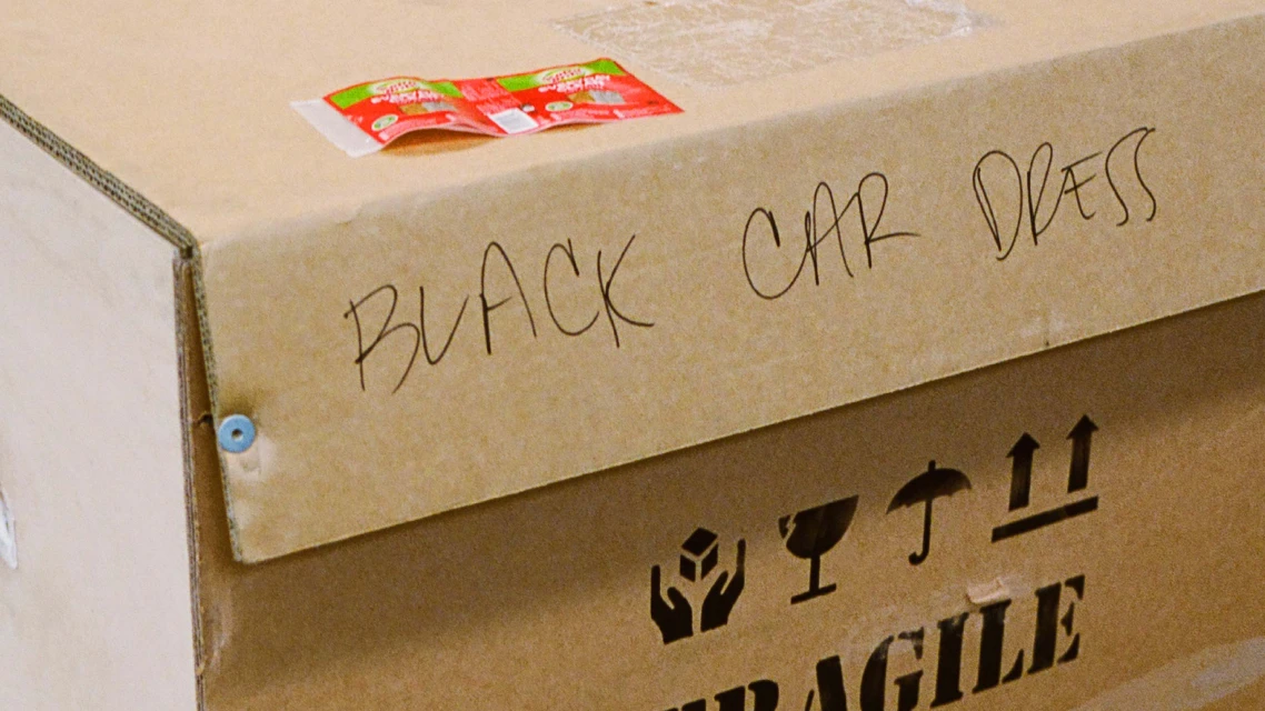 Close-up of a brown cardboard shipping box with handwritten text that says 'BLACK CAR DRESS' and package handling symbols.