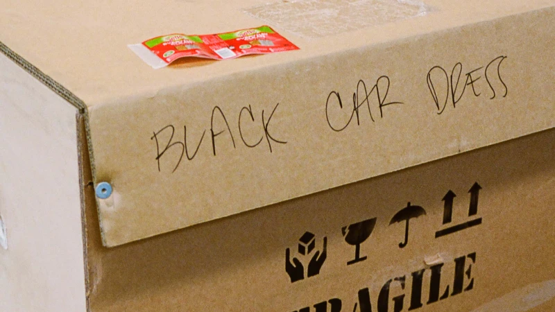 Close-up of a brown cardboard shipping box with handwritten text that says 'BLACK CAR DRESS' and package handling symbols.