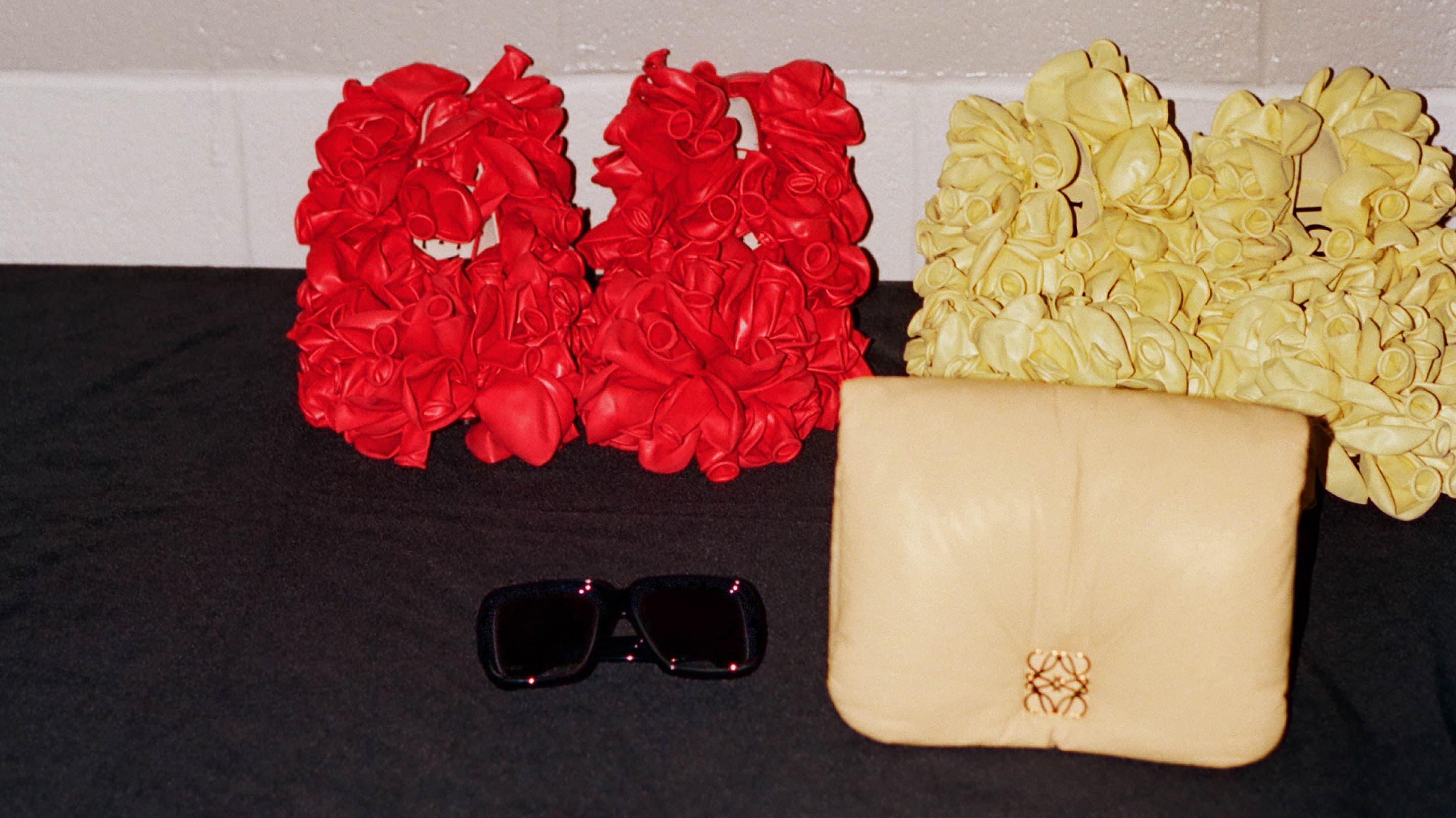 Still life of Loewe handbag, sunglasses, and puffy balloon-texture bags in red and beige on a black surface, indoors, simple studio shot.