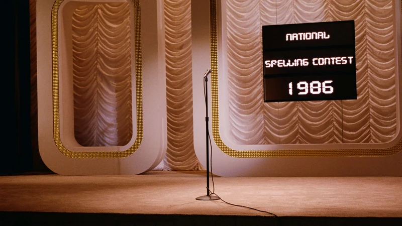 A stage with a microphone stands ready for the National Spelling Contest of 1986, lit by warm lighting. The backdrop has elegant curtains.