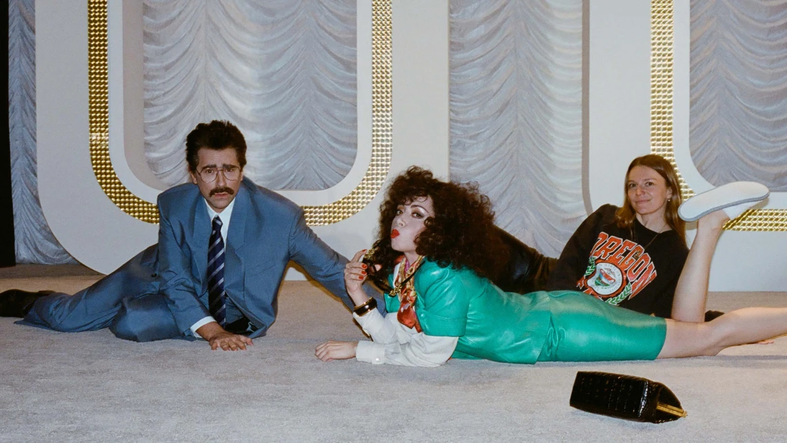 Three people pose on a grey carpet. A man in a blue suit, a woman in green leather dress, and another woman in a sweatshirt.