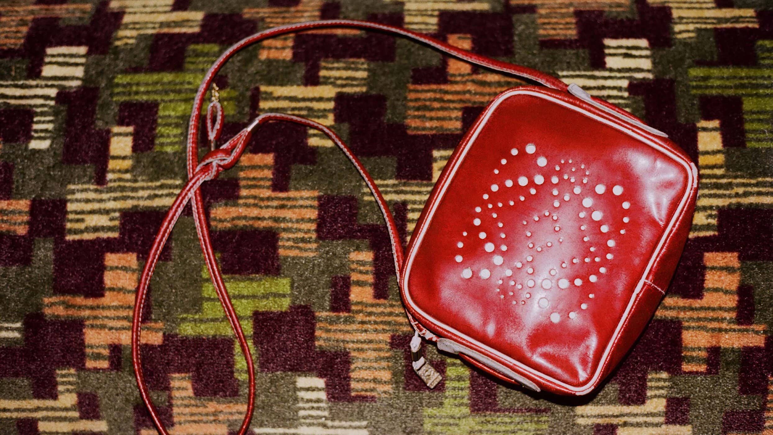 Red square purse with white circular pattern design and red and white strap over a brown, green, and beige patterned rug.