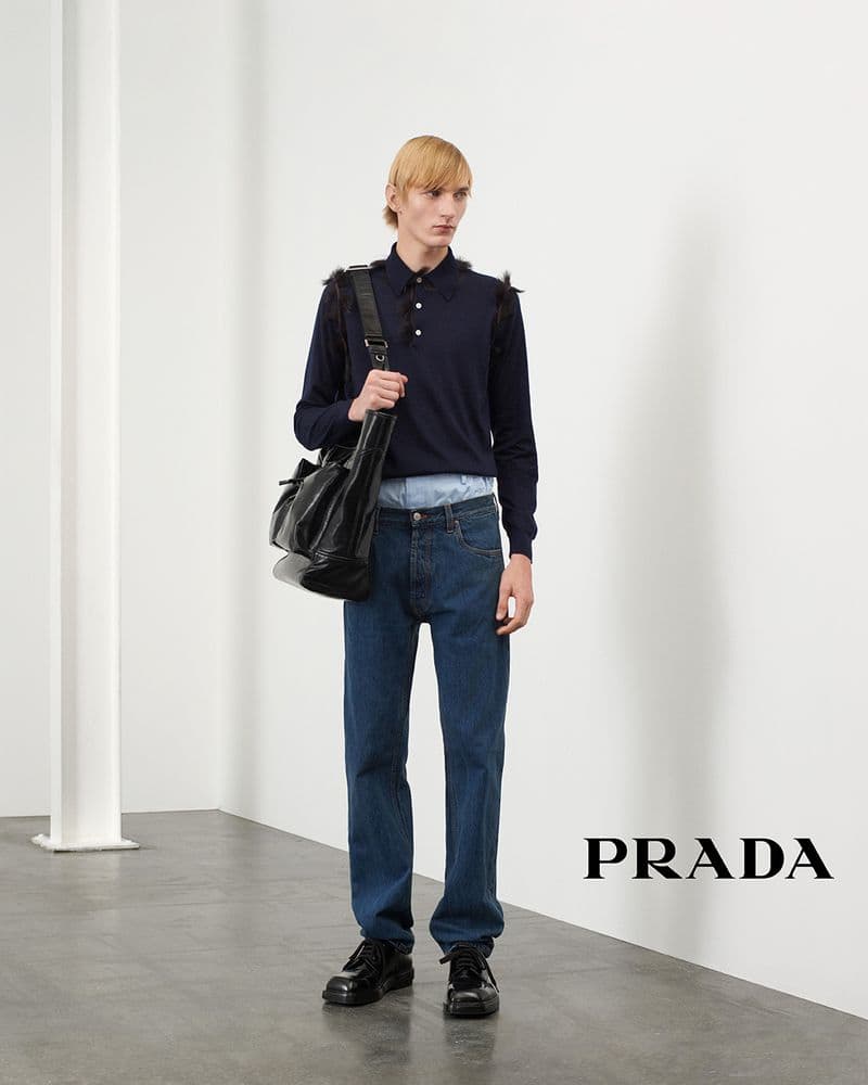 Blond male model wearing a dark blue long-sleeve polo shirt with fur trim and blue jeans, carrying a large black leather bag.