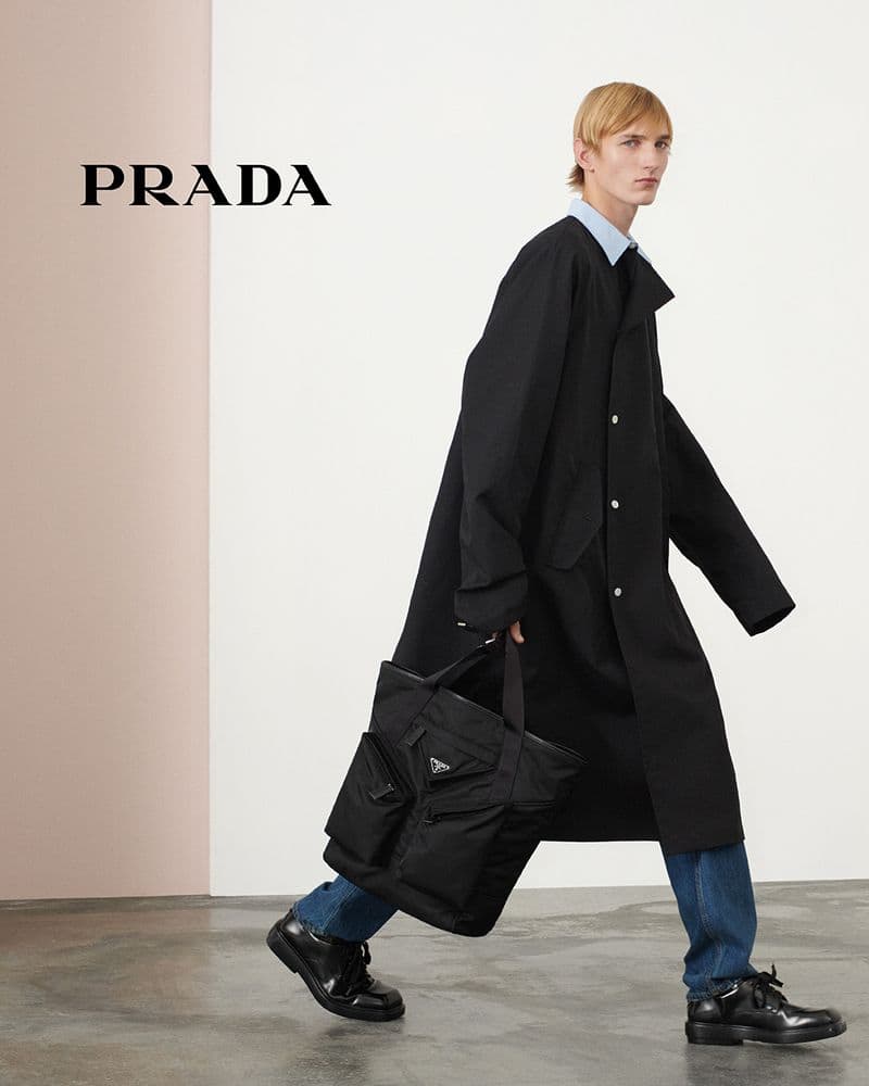 Fashion editorial of a man in a black coat carrying a black tote bag and walking on a gray floor against a two-tone gray wall.